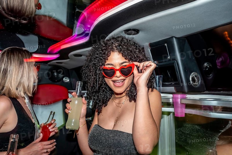 Young woman smiling and adjusting her heart shaped sunglasses, holding a drink bottle, while celebrating and having fun with friends inside a luxurious party limousine