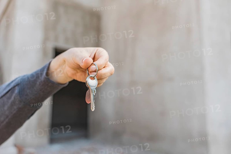 Close-up keys in hand. Woman in a hard hat and orange safety vest holds a set of keys in front of a construction site. The background shows unfinished buildings and debris