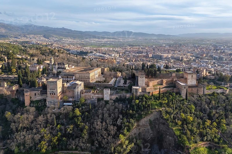 Wide aerial view of the Alhambra palace and fortress complex above Granada, Spain, surrounded by wooded hills with the Sierra Nevada on the horizon beneath a deep blue sky
