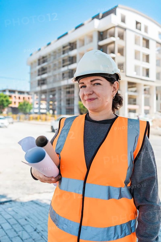 A young Hispanic woman wearing a hard hat and orange safety vest holds blueprints at a construction site with buildings in the background