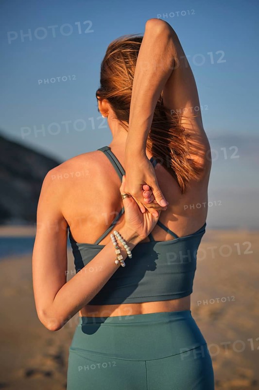 Athletic woman seen from behind holds Gomukhasana Cow Face Pose arms, clasping her hands behind her back during an outdoor yoga practice on a sunny sandy beach, featuring sand beach and stretching