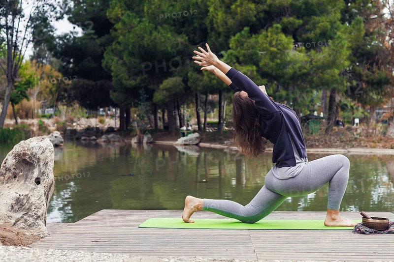 A young Hispanic woman practices yoga on a green mat by a serene pond surrounded by trees. She is in a lunging pose with arms extended