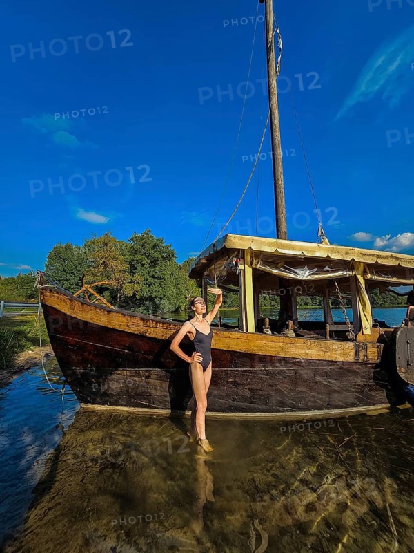 Wide angle view of a slender woman in a black swimsuit standing in a lake near an old wooden ship and covering her face from the sun with her palm, A woman stands by a wooden boat on a clear lake under a bright blue sky