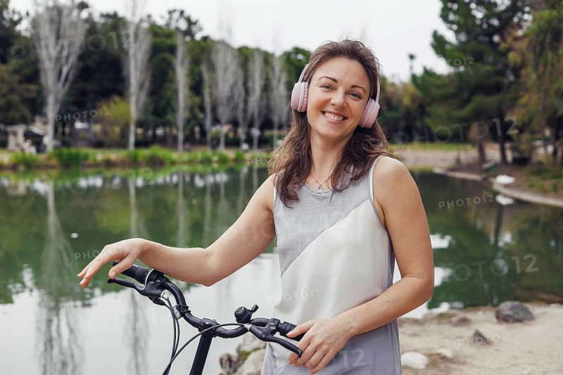 A young Caucasian woman with brown hair stands beside a bicycle near a serene lake. She wears pink headphones and smiles, enjoying the outdoors