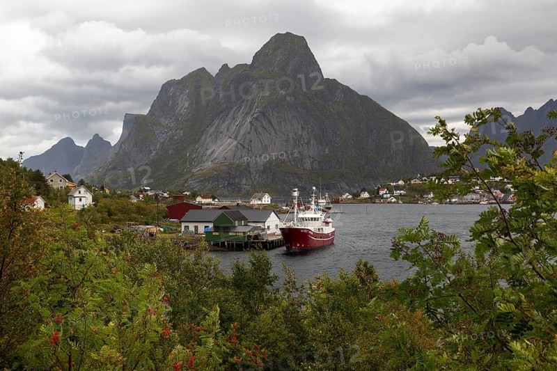 Red fishing boat on pier in Reine harbour in front of the majestic Olstinden mountain under cloudy sky, Reine, Lofoten, Norway