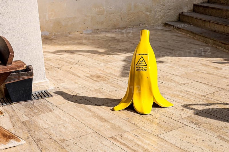 A yellow banana-shaped caution sign stands on a wooden floor. The sign warns of potential slipping hazards in a public area