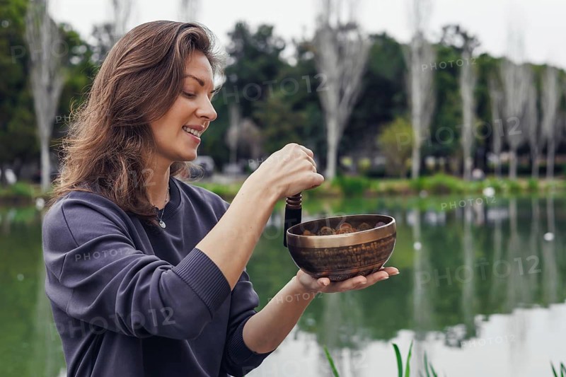 A young Caucasian woman with long brown hair holds a wooden bowl near a serene lake. Trees and greenery are visible in the background