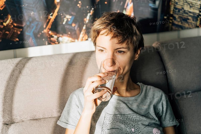 Portrait of a short-haired young woman drinking water from a glass, Woman sitting on a couch, drinking from a glass, with sunlight illuminating and a city background