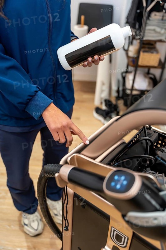 Professional beautician holding a product bottle and adjusting settings on a modern aesthetic device, preparing for a laser hair removal session in a clinic