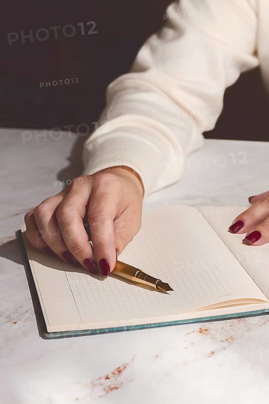 A person writes on an open notebook using a gold pen while sitting at a wooden table. The scene takes place in daylight. The focus is on the writing activity