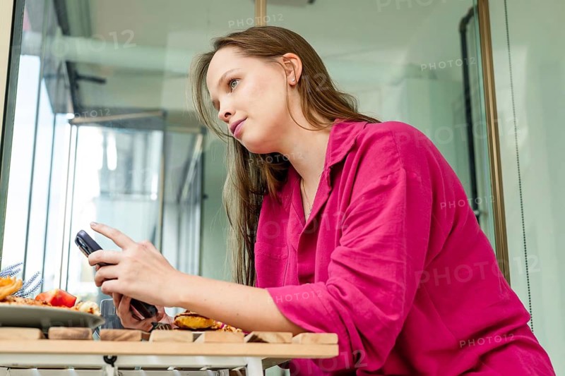 A young Caucasian woman with long brown hair sits at a table, using her phone. A plate of food with fruits and pastries is in front of her. Natural light fills the space