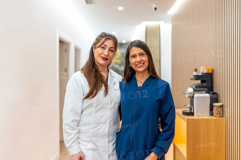 Two smiling female doctors, one wearing a white lab coat and the other a blue scrub, posing in a modern clinic or beauty salon hallway, providing services for health and wellness