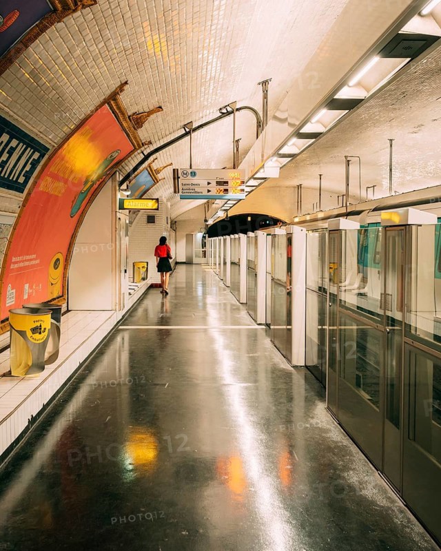 Underground platform of a subway station with a vaulted, white-tiled ceiling, large advertising posters on the left wall and a waiting person, on the right, glass platform doors delimit the track area with reflecting, dark flooring. Paris, Paris Metro, Ile-de-France, France