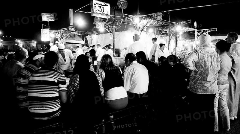 Black and white photo, people sitting at food stalls at night, market stalls in the background, evening food stalls at Djemaa el Fna, Marrakech, Morocco
