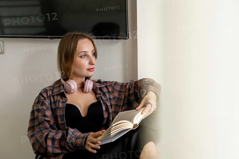 Young Caucasian woman with long brown hair, wearing headphones, reading a book while sitting on a chair in a modern indoor setting