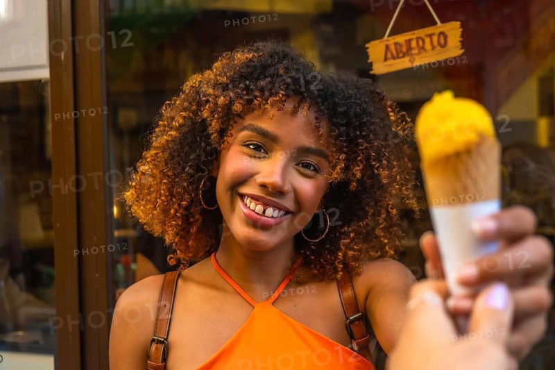 Young happy woman with afro textured hair smiling cheerfully, holding out a delicious mango ice cream cone on a warm urban day, embodying joy and summer vibes