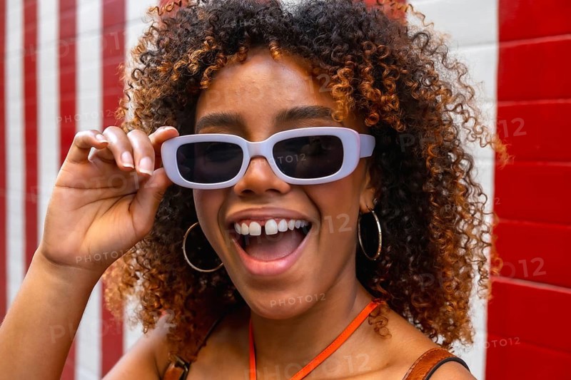 Young woman with curly hair and sunglasses smiling at camera, showing gap toothed grin and joyful, confident expression in vibrant urban summer portrait with red striped background