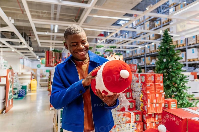 A young Black man smiles while holding a large red Santa toy in a festive store filled with Christmas decorations and gifts