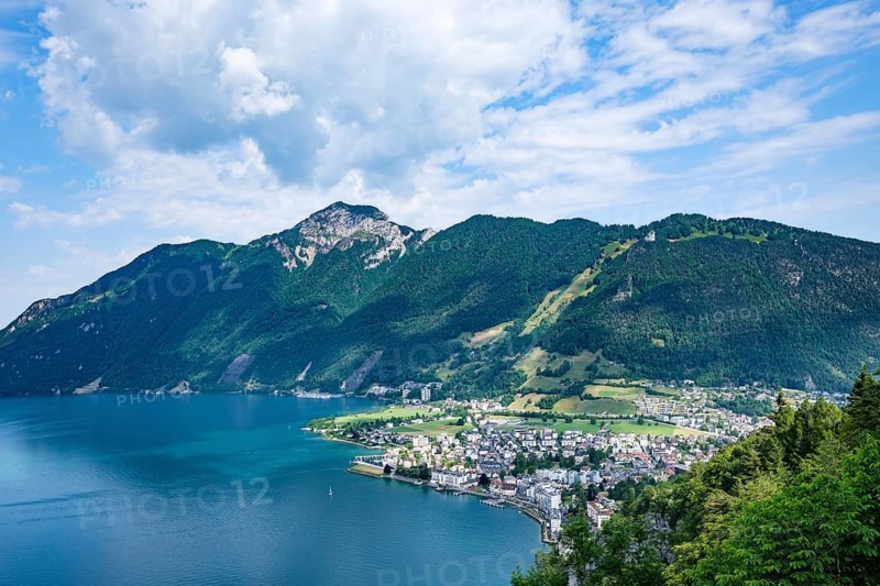 Urban coastal community at the foot of a mountain with houses, blue water and wooded hills under a cloudy sky, Brunnen, Canton of Schwyz, Switzerland