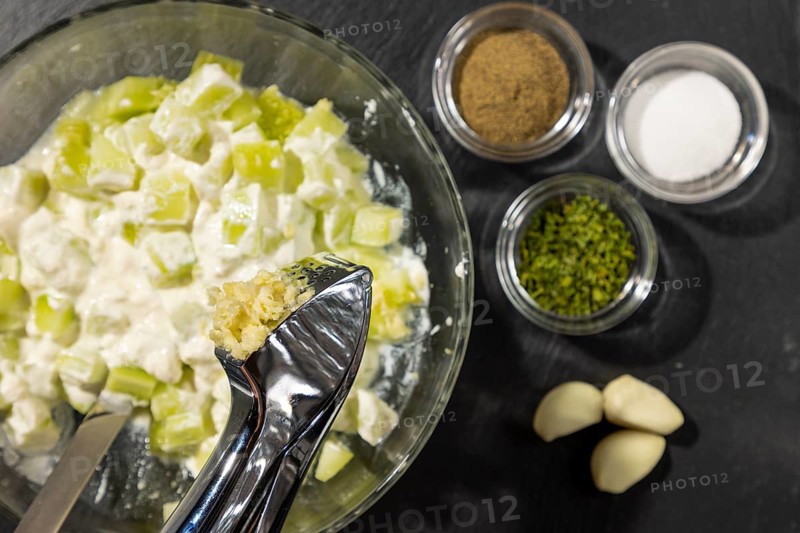 Garlic is pressed into sliced cucumber and sour cream in a bowl, plus pepper, salt and chives