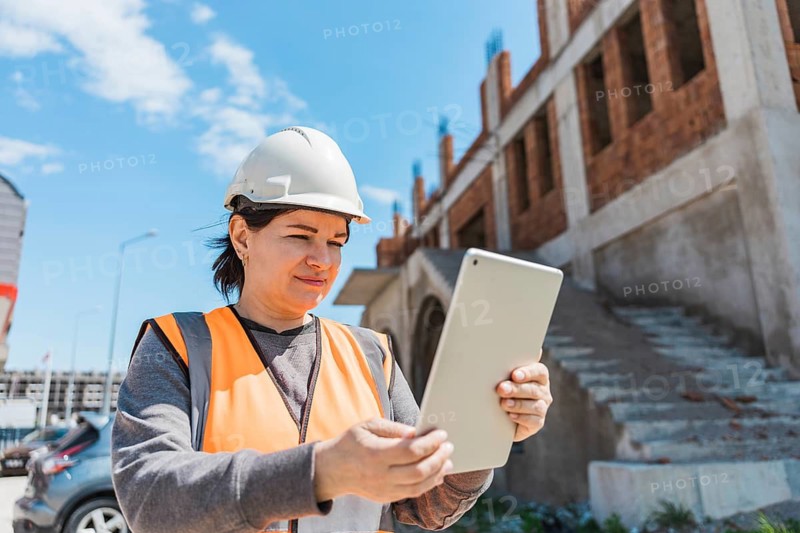 A young Hispanic woman in a hard hat and orange vest uses a tablet at a construction site. The background features a partially built structure