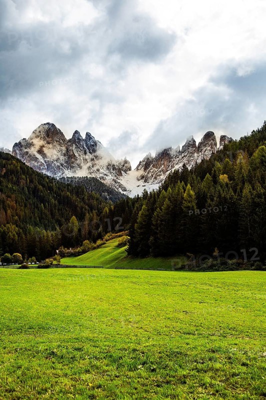 Wide green meadow in front of a monumental mountain chain, partly covered with snow, under a dramatic sky, view of the Geisler Group mountain from the Funes valley. Funes, Trentino Alto Adige, Italy