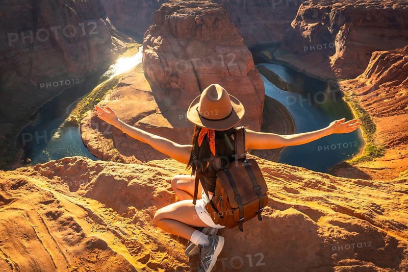 Young woman hiker sitting on a sandstone cliff, arms outstretched, feeling freedom and happiness while admiring the dramatic landscape of horseshoe bend and the colorado river