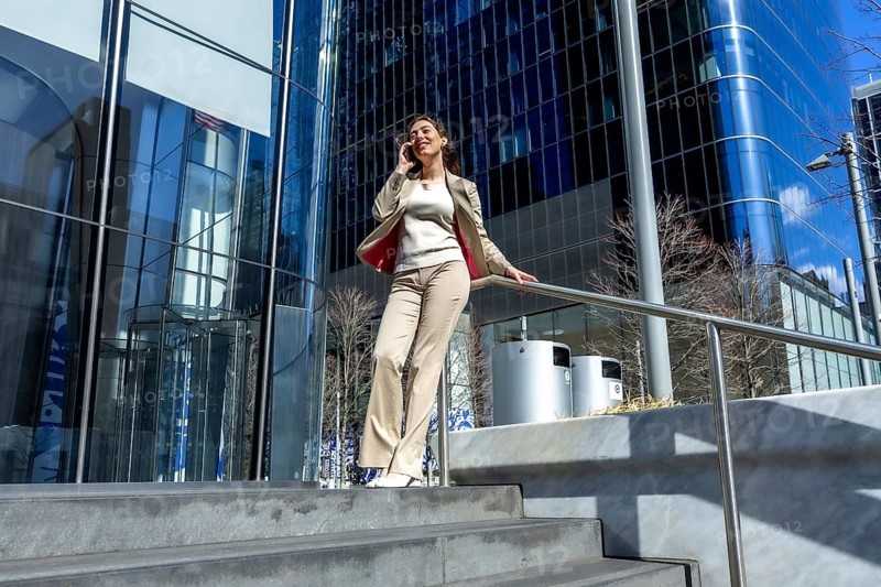 Confident businesswoman on steps talking on a smartphone with glass office towers reflecting the city skyline, conveying urban career success, networking and leadership