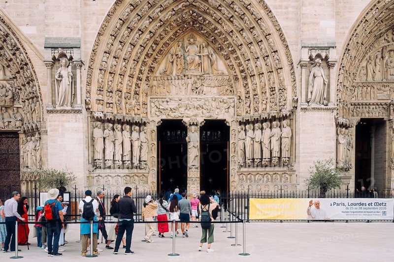 Central portal of the western façade of the Gothic Notre-Dame de Paris cathedral, known as the Portal of the Last Judgment, with richly decorated rows of sculptures in the archivolts and the detailed tympanum relief above the open wooden entrance doors, in front of which tourists and visitors queue at a barrier and hangs in the background on the right a horizontal information banner about a Pope's trip. Paris, Ile-de-France, France