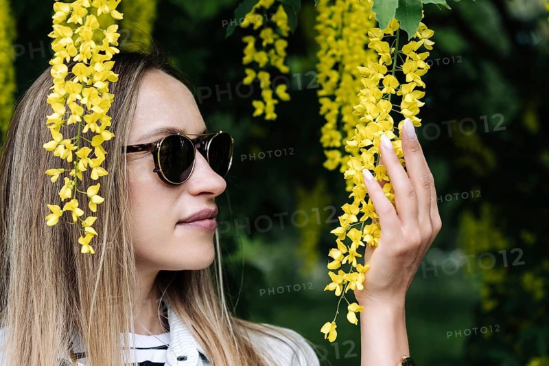 A woman holds in her hand the inflorescence of Laburnum anagyroides, A woman wearing sunglasses admires hanging yellow flowers in a lush outdoor setting