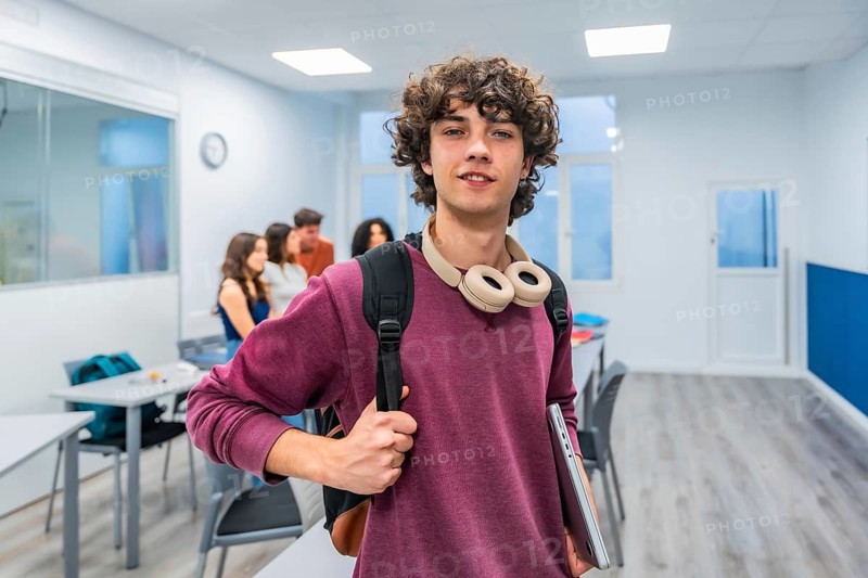 Young male student with curly hair and headphones around his neck holds a laptop and backpack, smiling confidently in a classroom while peers chat in the background
