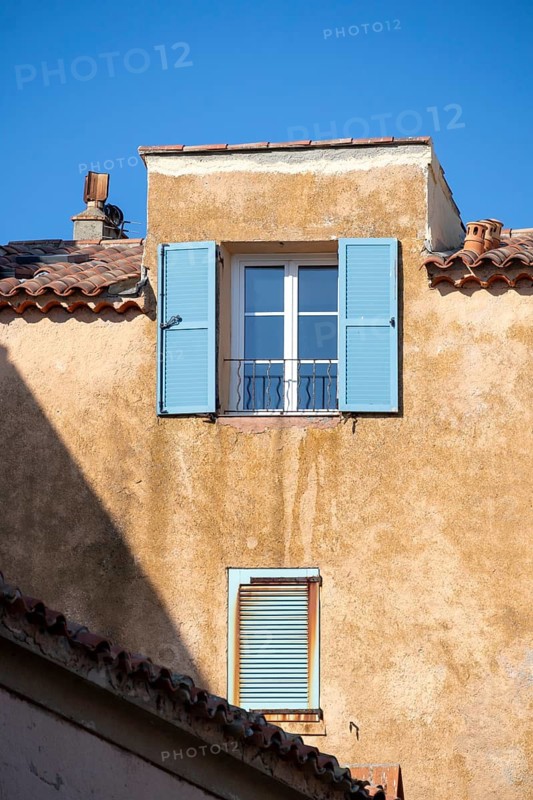 Blue shutters and a small window stand out on a weathered ochre facade beneath terracotta roof tiles, Saint-Tropez, Var, Provence-Alpes-Cote d'Azur, France