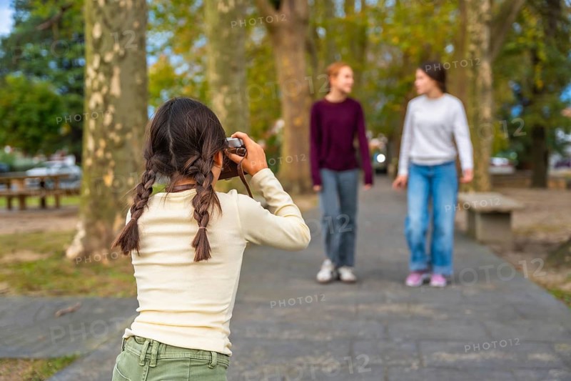Young girl holding a vintage camera, looking through the viewfinder and capturing casual moments of two blurred friends walking and chatting happily along a path in a park on a sunny day