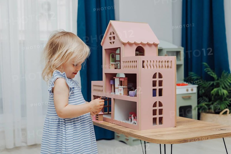 Focused child in a blue dress plays with a dollhouse on a wooden table inside a room