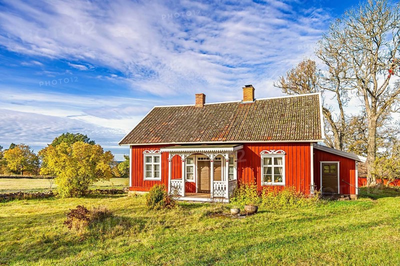 Red wooden cottage with white trim stands in a rural landscape surrounded by autumn trees. Bright sunlight and open fields highlight the charm of Swedish countryside architecture, Sweden