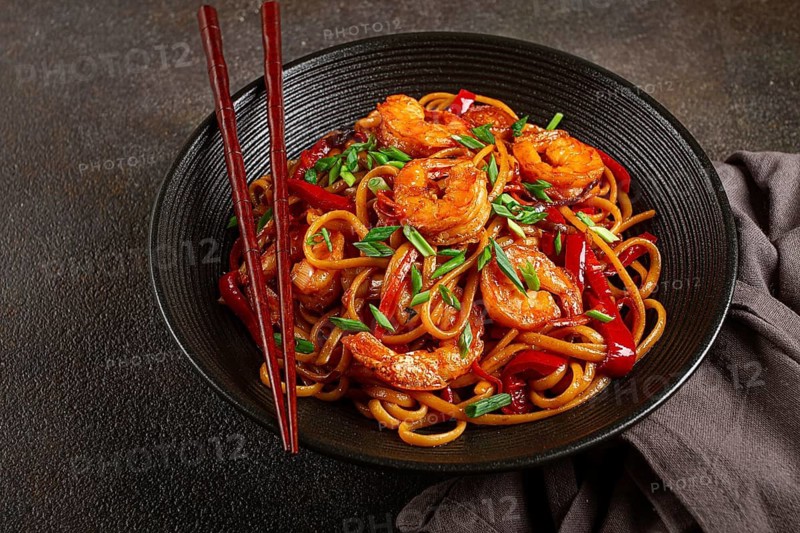 Udon stir-fry shrimp noodles with red bell pepper and green onion, served in a black bowl, with chopsticks, dark background