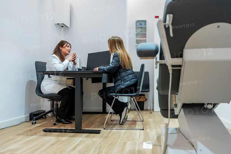 Female doctor in a professional white coat discussing treatment options with a young woman, sitting together at a desk in a modern aesthetic clinic for wellness and personal care