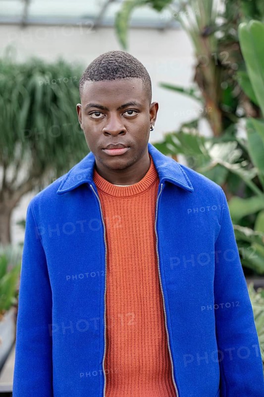 A young African man stands in a lush green environment. He has short black hair and wears a blue jacket over an orange sweater