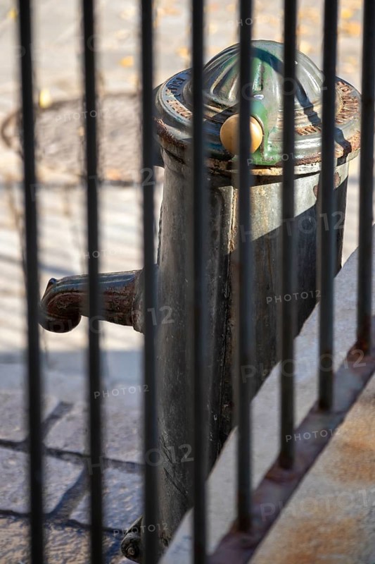 A green metal street fountain is seen through vertical black railings on stone paving, Saint-Tropez, Var, Provence-Alpes-Cote d'Azur, France