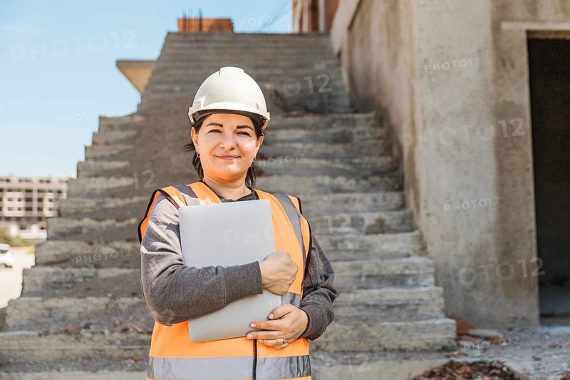 Woman stands on construction stairs wearing a hard hat and safety vest, holding a laptop. The construction site is visible in the background