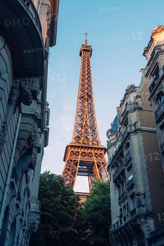 Eiffel Tower in Paris between historic Haussmann-style residential buildings and green treetops against slightly cloudy blue sky, Paris, Île-de-France, France