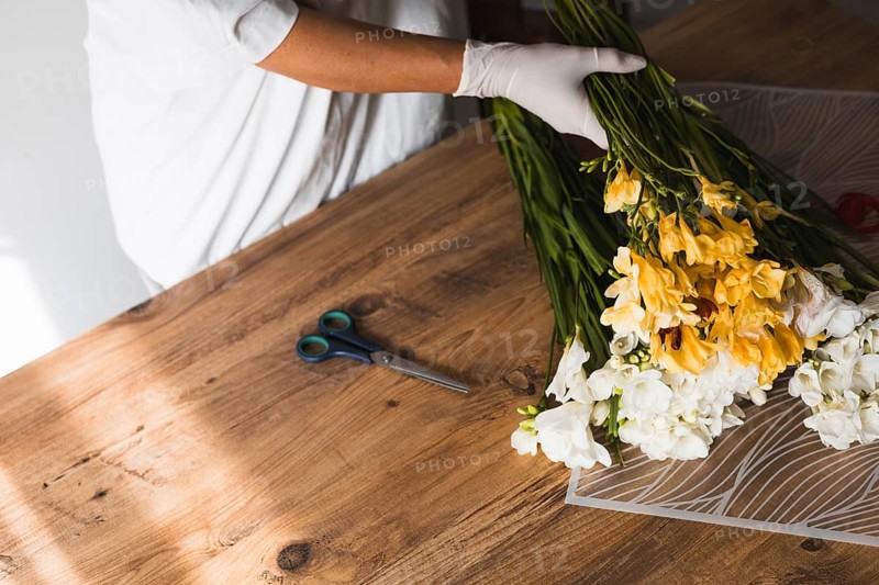 A florist in gloves is carefully arranging fresh flowers on a wooden table. The workspace is bright, showcasing vibrant blooms being prepared for a bouquet