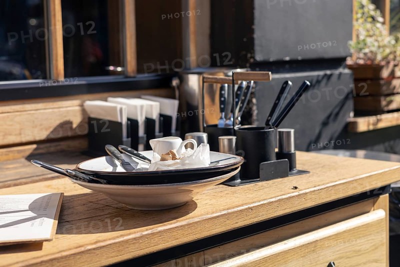 Dirty dishes on a table after client in restaurant. An inviting outdoor cafe, wide variety of knives, a small cup, and utensils on a wooden table, warm and welcoming atmosphere for patrons