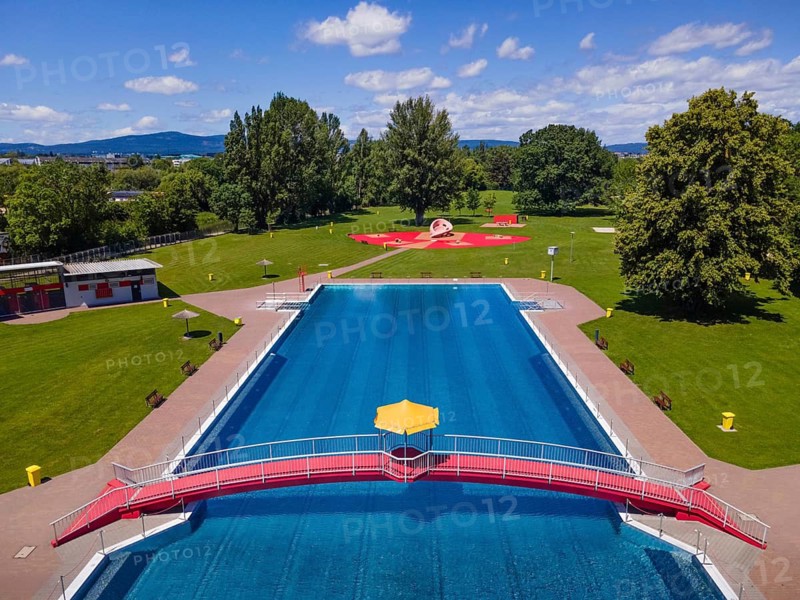 On a red bridge spanning a swimming pool at an open-air lido in Frankfurt, there is a yellow parasol in the centre. The deserted pool below is still waiting for the rush of bathers. The start of the swimming season is just around the corner. (Aerial view taken by drone), Eschersheim, Frankfurt am Main, Hesse, Germany
