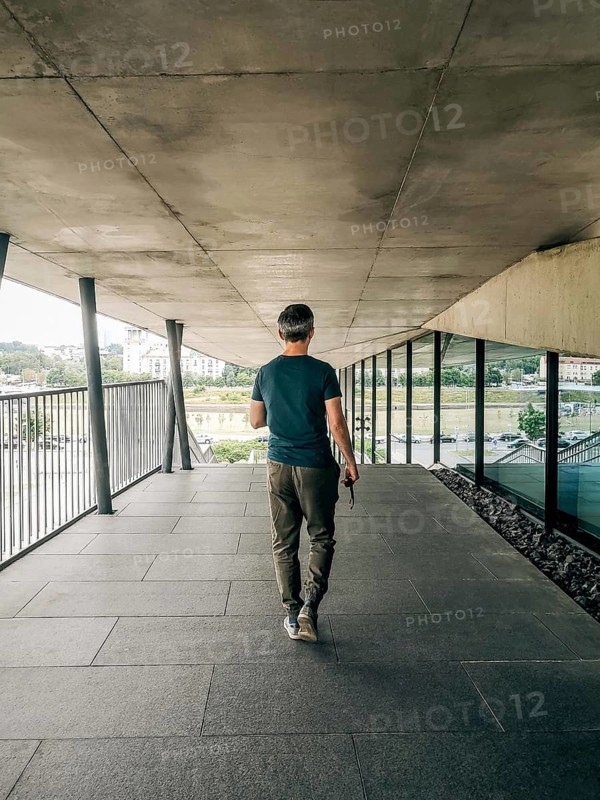 The view from behind walking a man in casual clothes, A man in casual attire walks down a modern, concrete corridor with an urban backdrop
