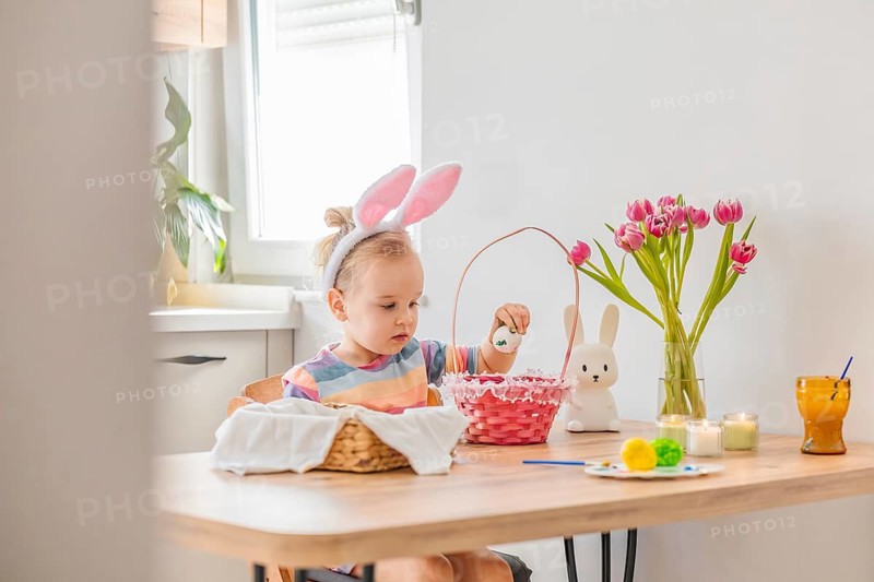 A young child wears bunny ears and dips an Easter egg into a basket at a table. Flowers and candles are around, creating a fun atmosphere for holiday crafts