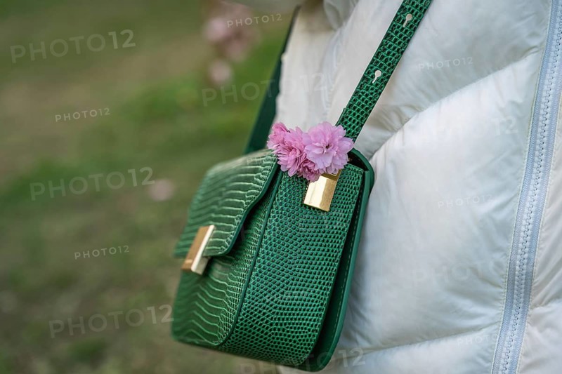 Women's green crossbody bag with pink japanese cherry blossoms (Prunus serrulata) Kanzan on the shoulder of woman in white jacket while walking in spring park, A green faux crocodile handbag with gold accents is adorned with a pink flower and draped against a white coat