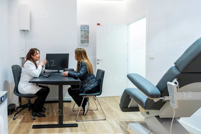 Female doctor speaking with a young woman about potential cosmetic procedures or laser hair removal, sitting at a desk in a modern aesthetic clinic with a treatment chair visible