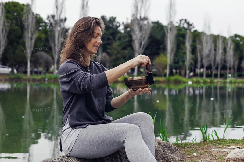 A young Caucasian woman with brown hair sits by a lake, holding a bowl. She wears a gray sweater and light gray pants. The background features trees and water
