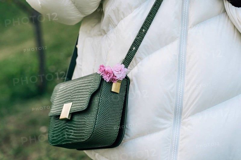 Fashion Stylish Reptile Imitation Women's Handbags With Pink Japanese Cherry Blossoms (Prunus serrulata) Kanzan On Woman's Shoulder While Walking In Spring Garden, A green textured handbag adorned with pink flowers hangs on a person in a white coat outdoors